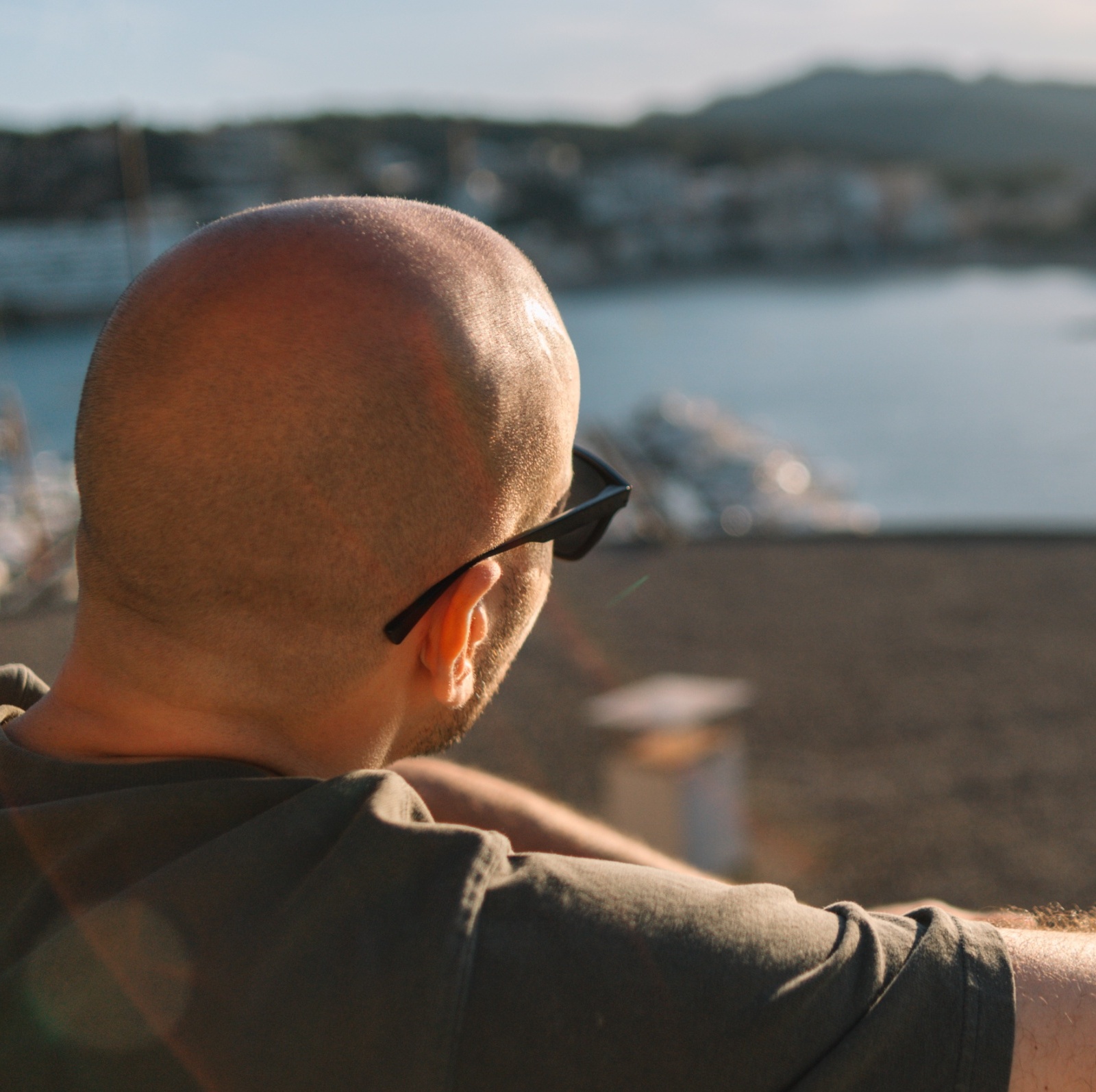 A bald man in glasses sitting at the beach overlooking a lake.