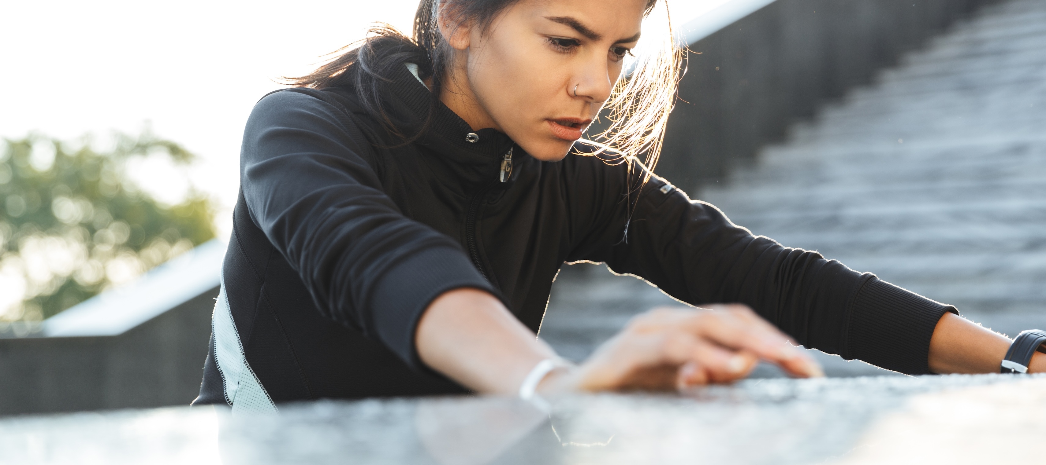 An athletic-looking young woman wearing a black jacket leans forward, hands resting on a grey surface. She appears tired.