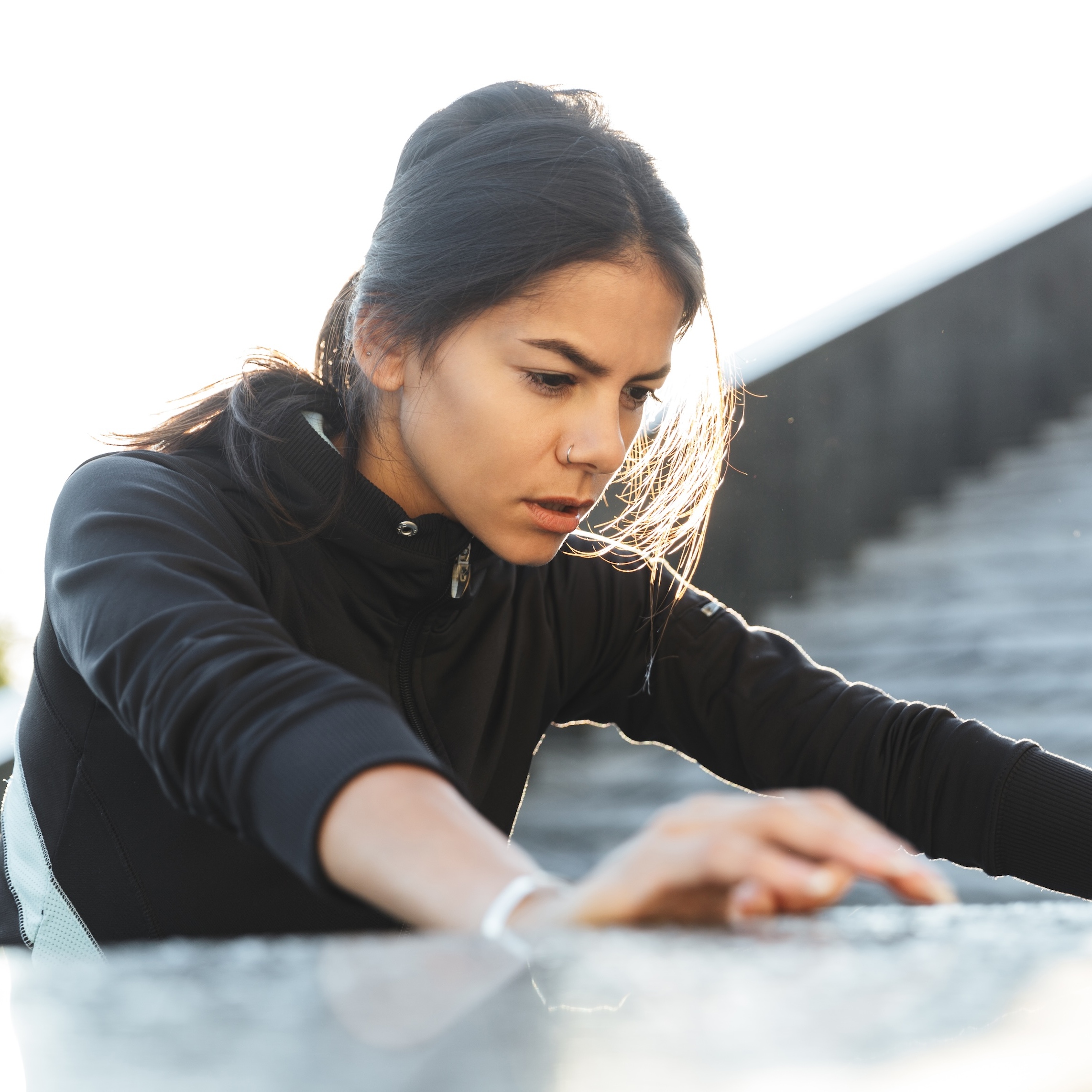 An athletic-looking young woman wearing a black jacket leans forward, hands resting on a grey surface. She appears tired.