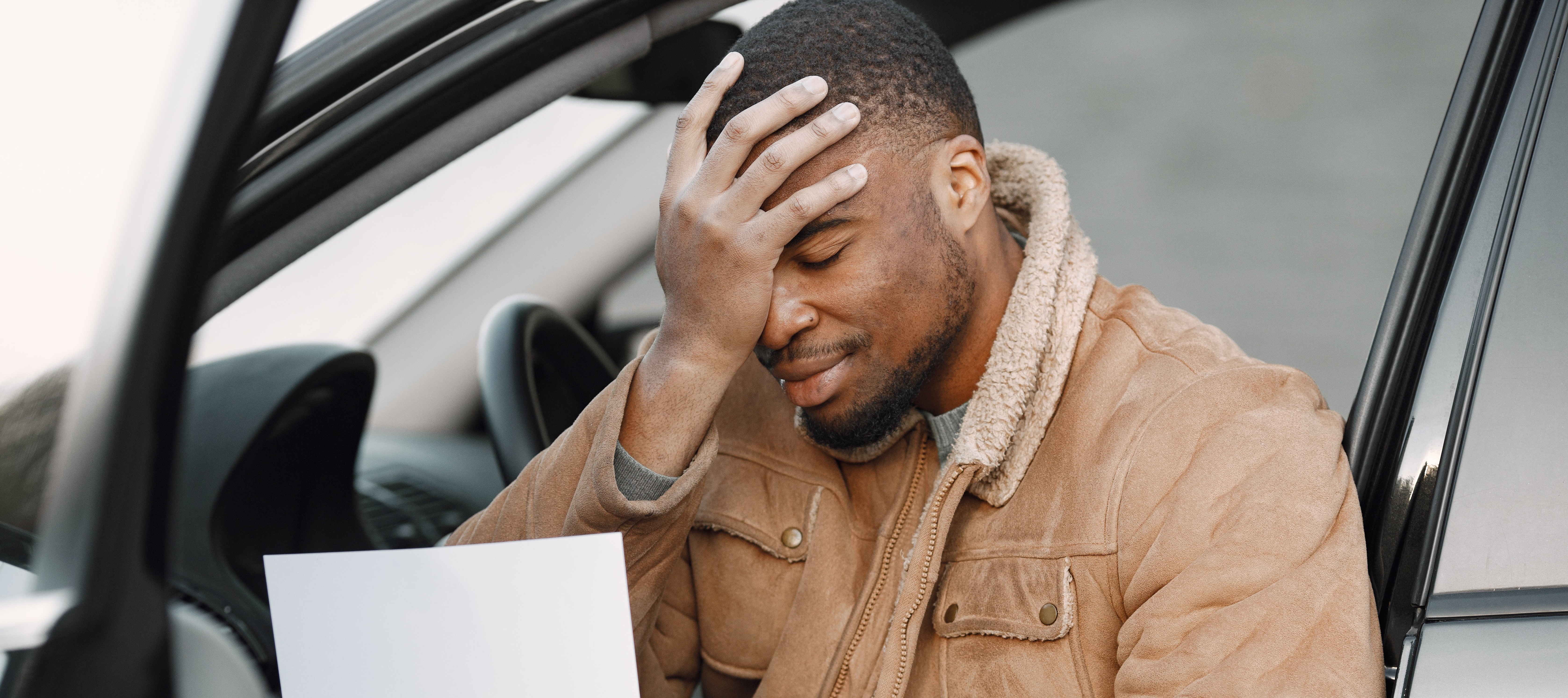 An adult man covers his face with his hand while sitting in the driver's seat of a car and holding a piece of paper.