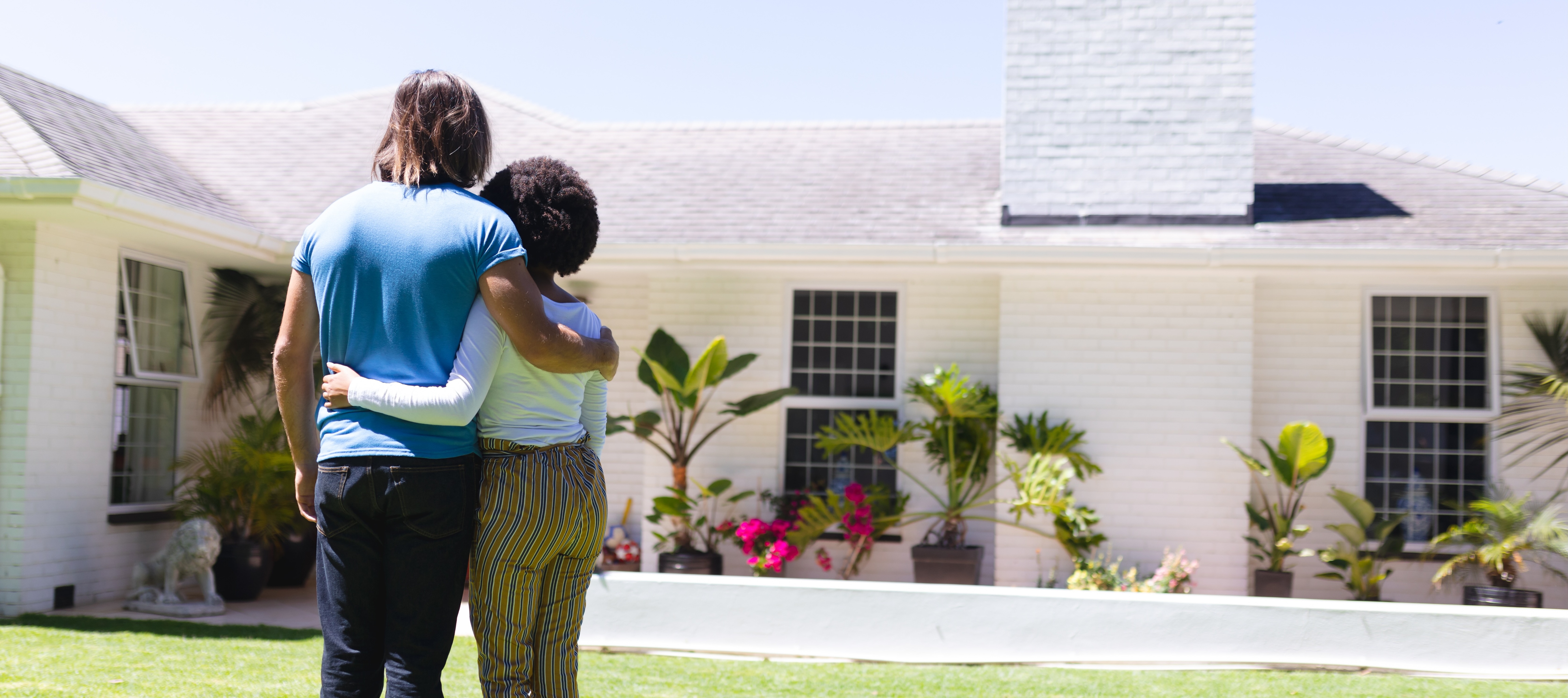 A couple stands embracing on a grassy lawn in front of a white, suburban home, their backs to the camera.