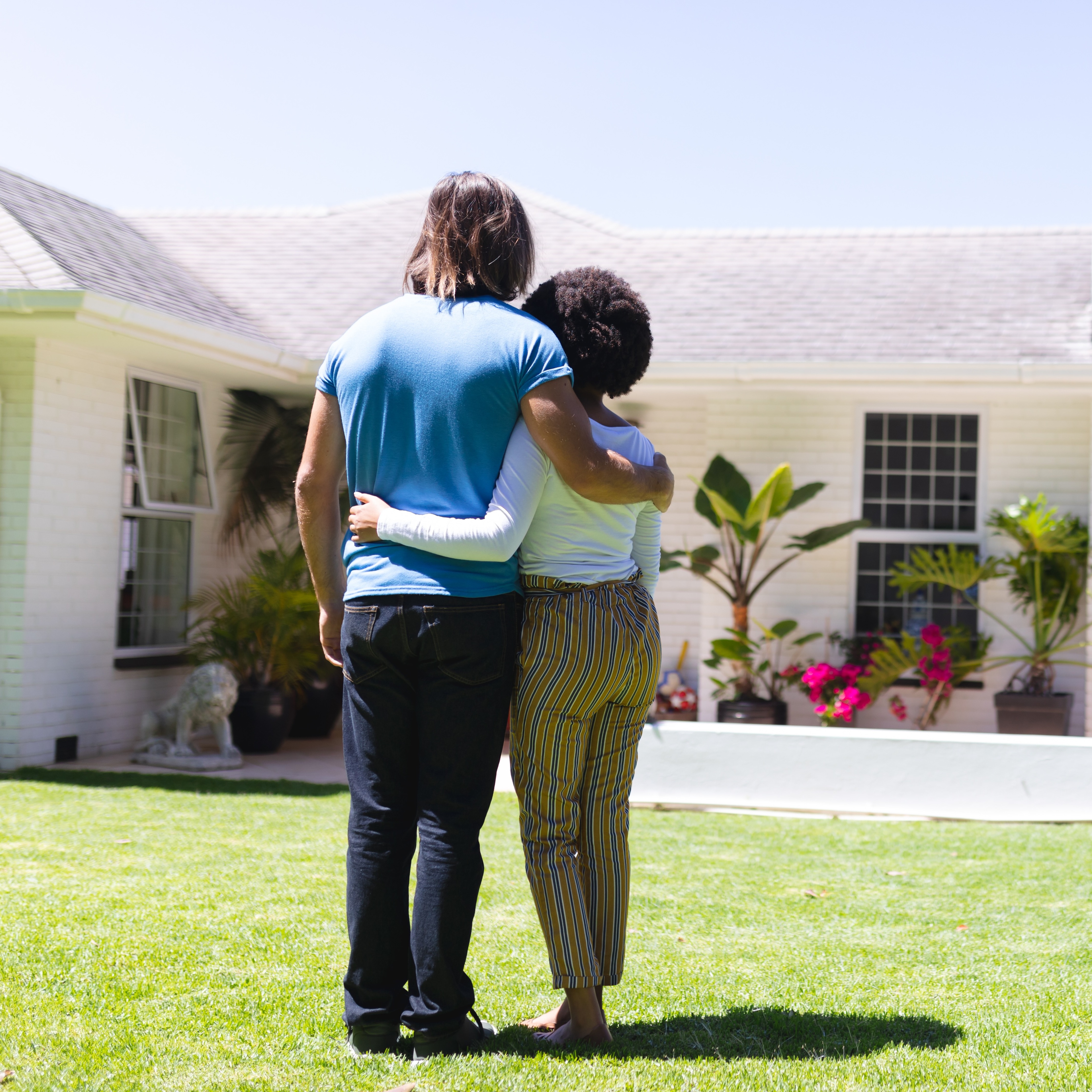 A couple stands embracing on a grassy lawn in front of a white, suburban home, their backs to the camera.