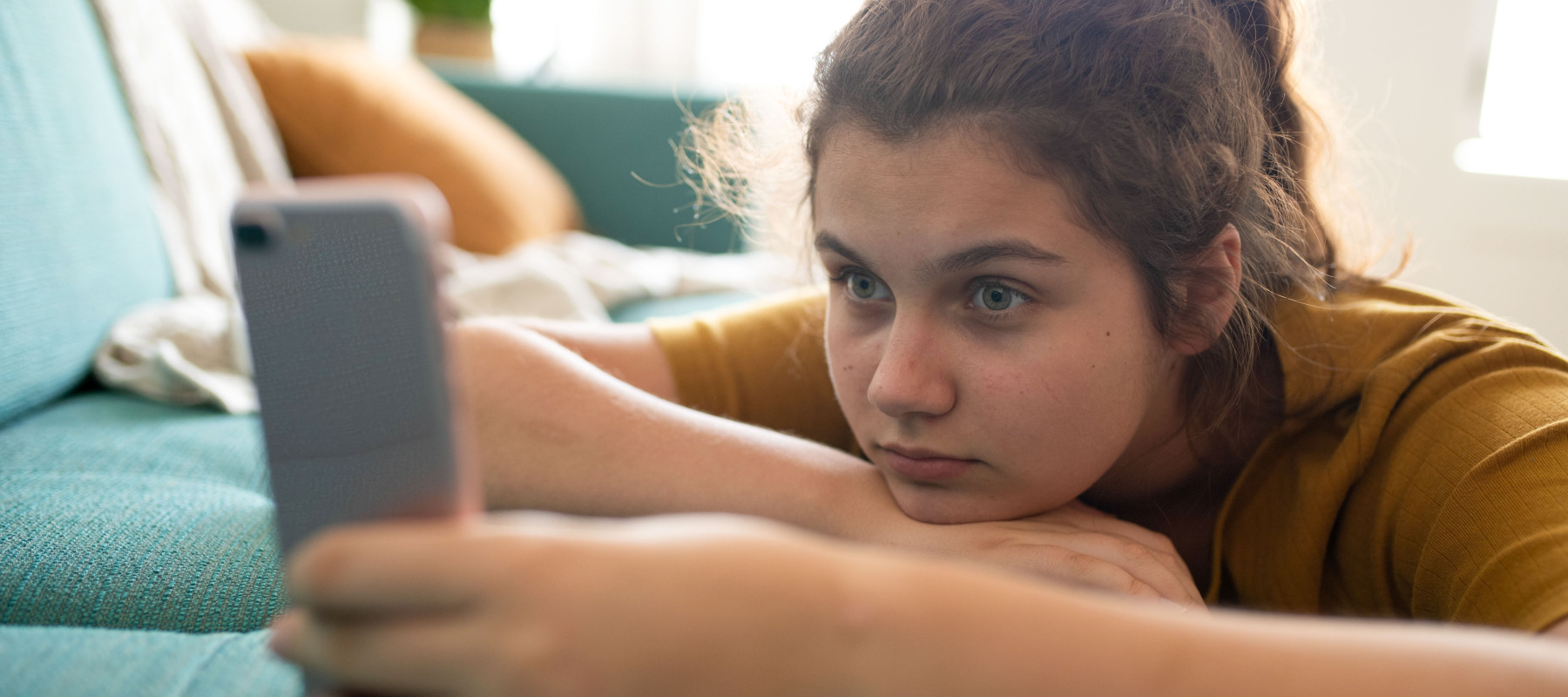 A young girl lays on a couch, holding and staring intensely into a phone screen.