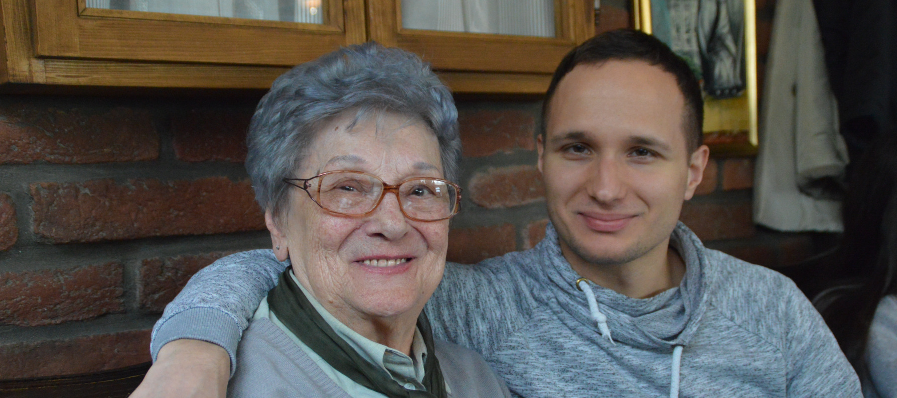 Older woman sits with young man, his arm draped over her shoulder. They're smiling.