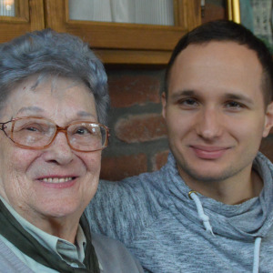 Older woman sits with young man, his arm draped over her shoulder. They're smiling.