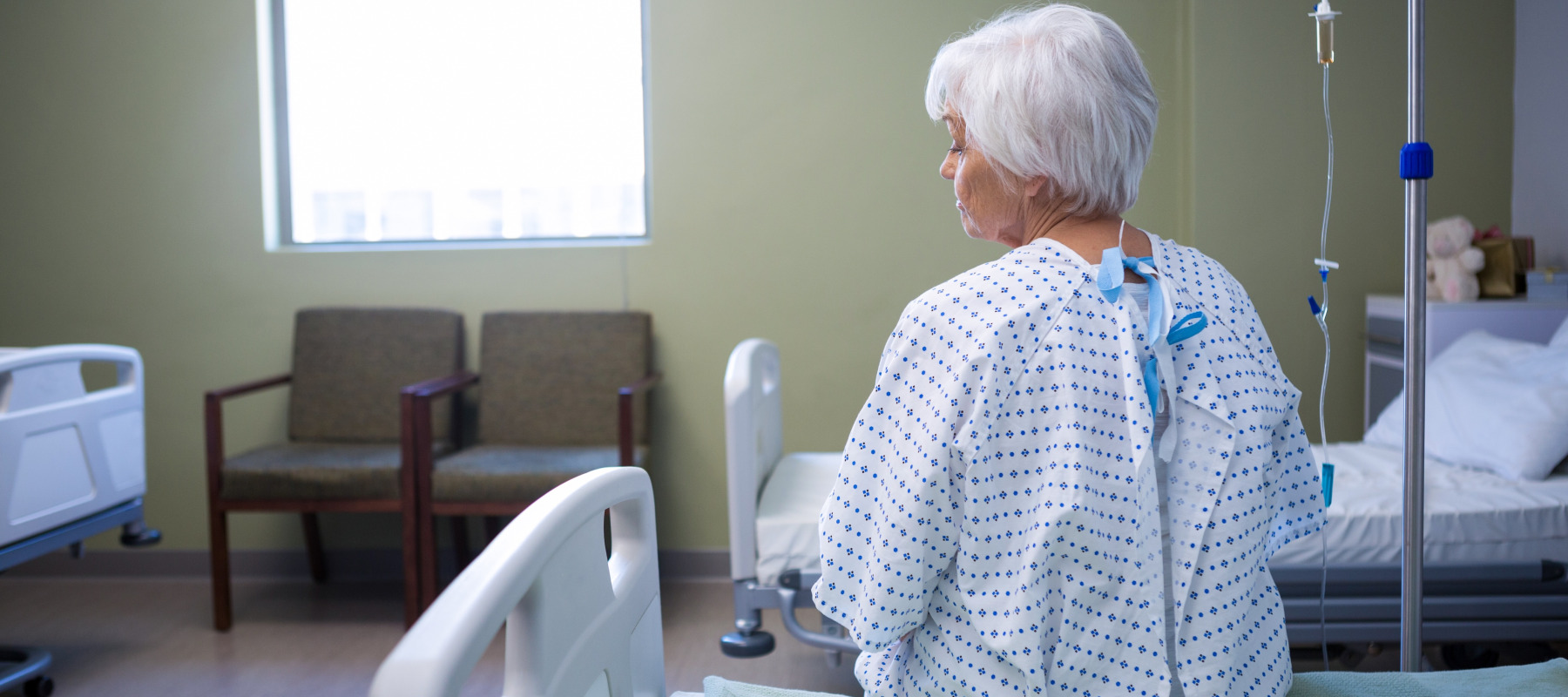 A senior woman with short, white hair sits on a hospital bed in a bright room.
