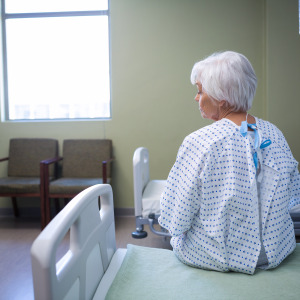 A senior woman with short, white hair sits on a hospital bed in a bright room.