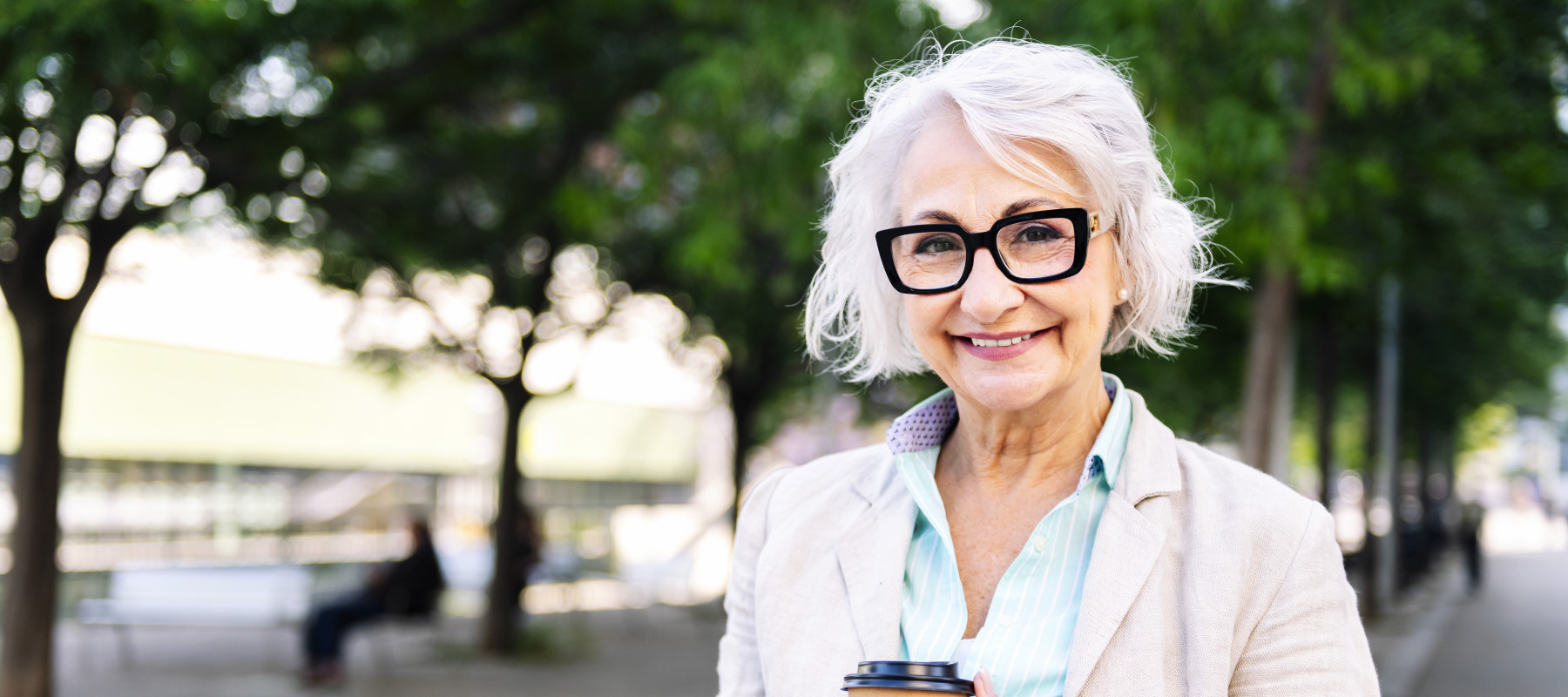A stylish older woman with short, wavy, white hair smiles at the camera, holding a coffee cup while standing in a city park setting.