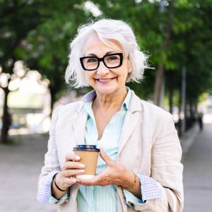 A stylish older woman with short, wavy, white hair smiles at the camera, holding a coffee cup while standing in a city park setting.