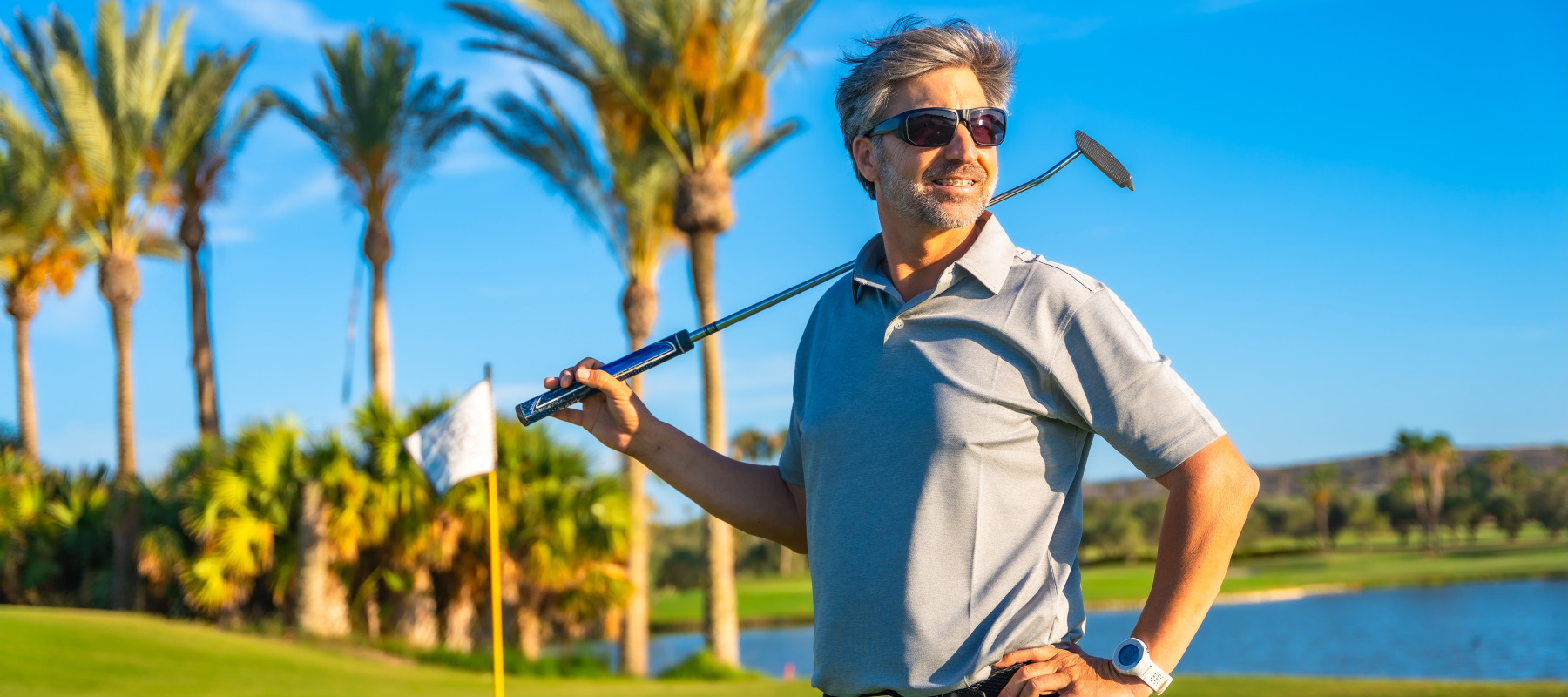 A smiling man on a brightly lit golf course stands with a golf club held over his shoulders.