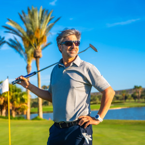 A smiling man on a brightly lit golf course stands with a golf club held over his shoulders.