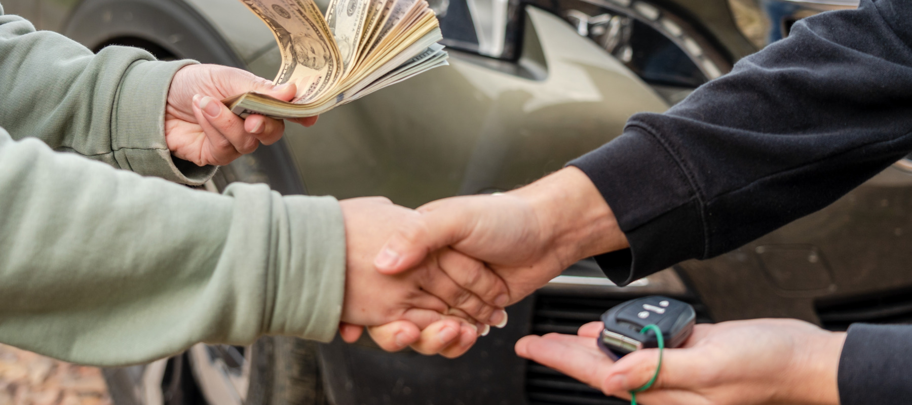 Hands exchange a wad of cash for a pair of car keys in a parking lot.
