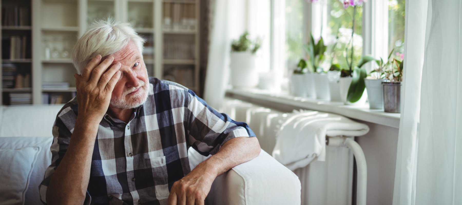 A senior man with gray hair sits on a white couch in front of a window.