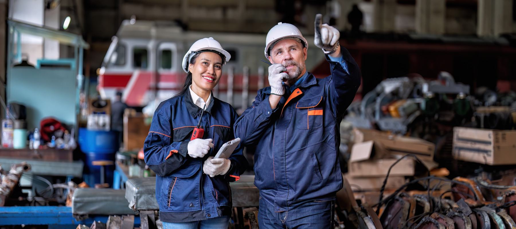 Two workers wearing hard hats and blue work uniforms stand in a manufacturing plant.
