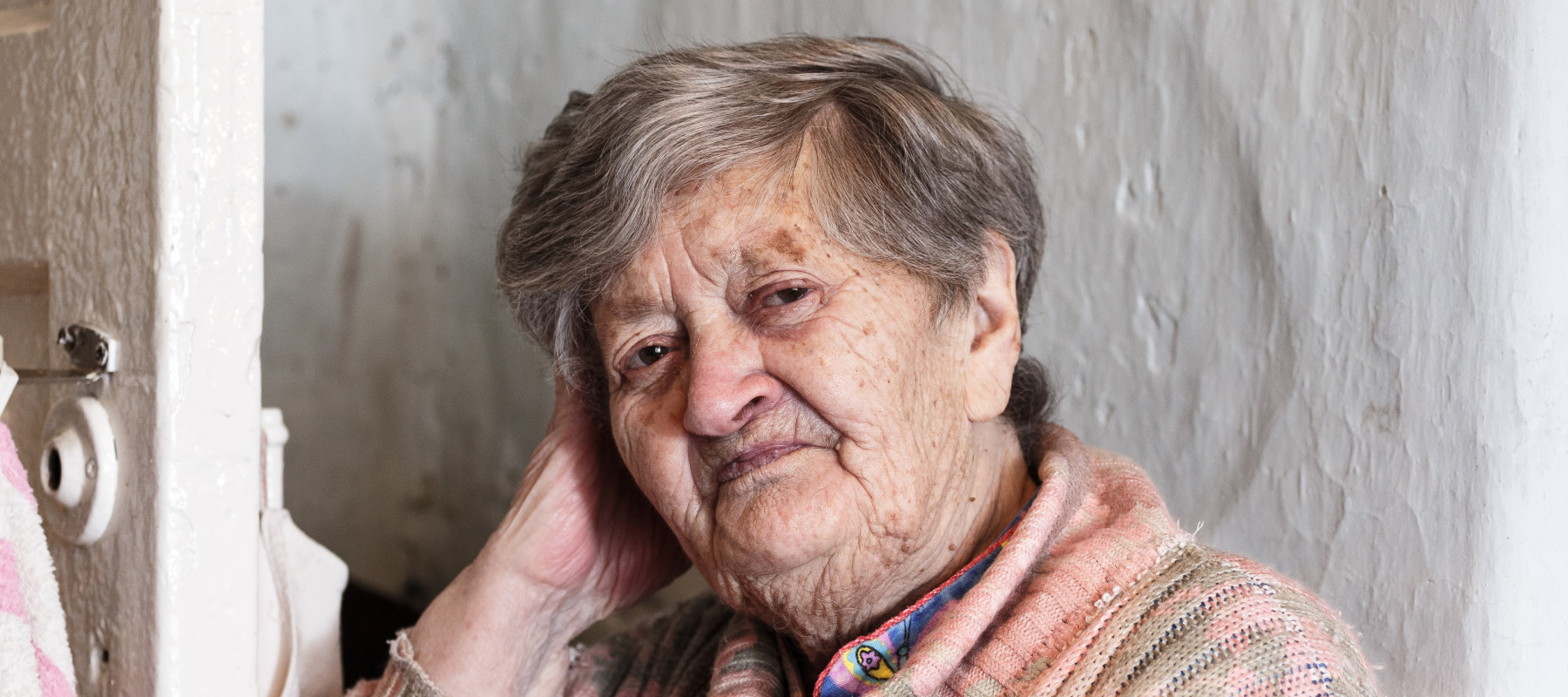 An older woman in a pink sweater looks at the camera, resigned and alone.