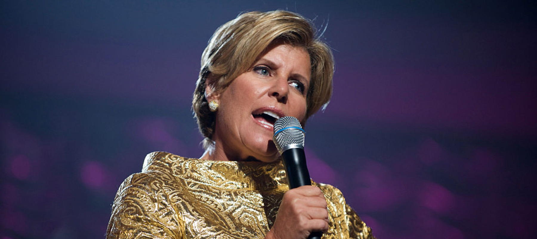 Suze Orman speaks into a microphone, looking disapproving and stern.
