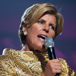 Suze Orman speaks into a microphone, looking disapproving and stern.