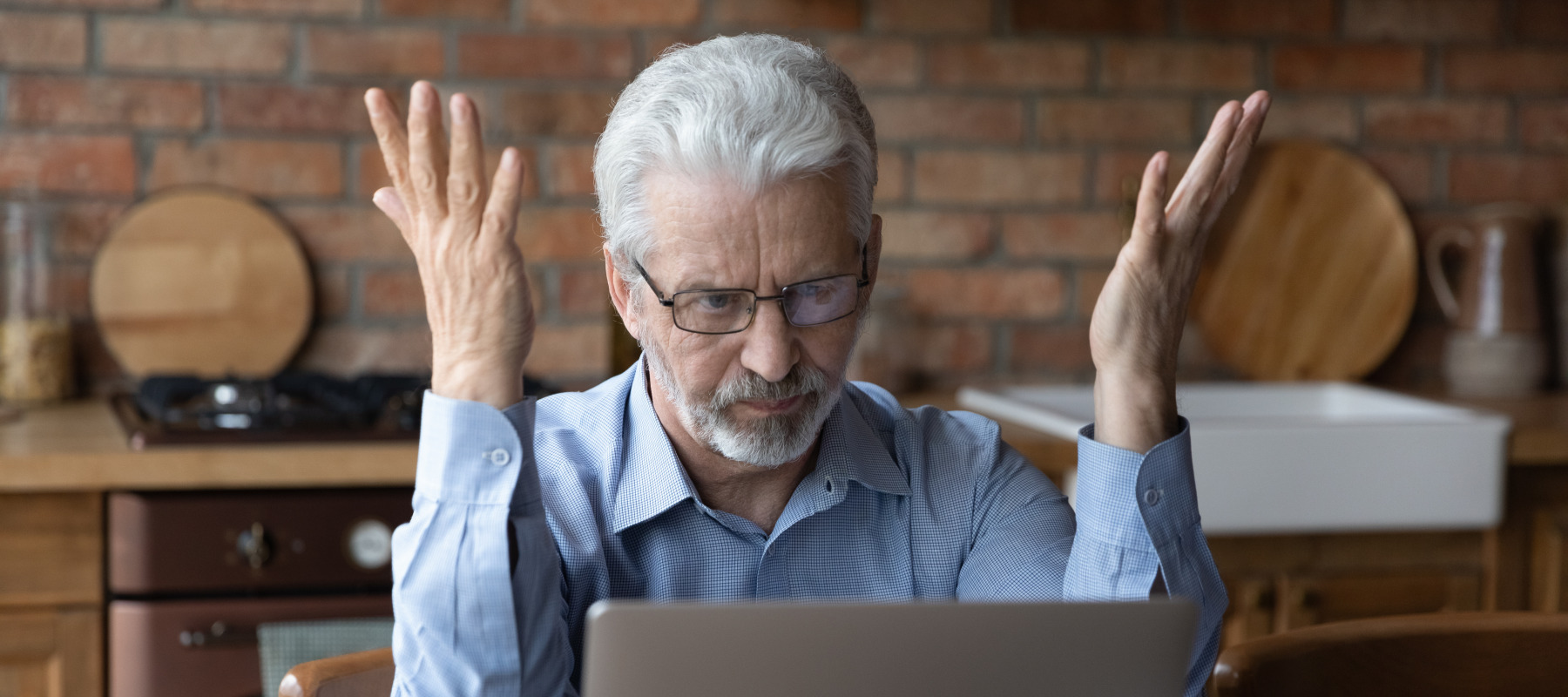 Annoyed worried elderly man sits in front of a laptop at a kitchen table.