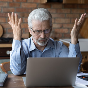 Annoyed worried elderly man sits in front of a laptop at a kitchen table.
