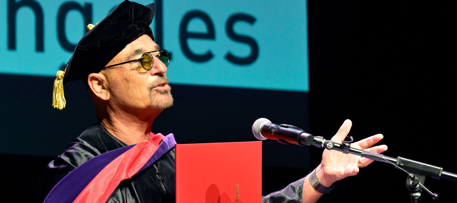 Howie Mandel gestures outwards as he delivers a speech on stage wearing graduation robes.