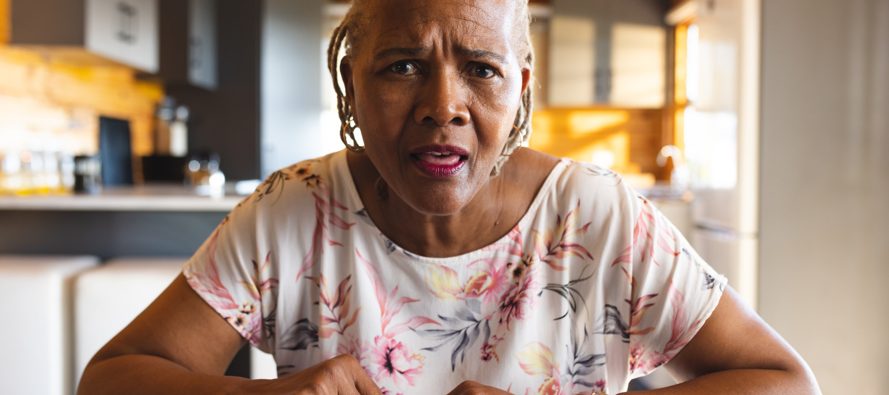 A woman with gray braided hair wearing a patterned blouse is shown indoors at a wooden table, looking at some kind of financial report.