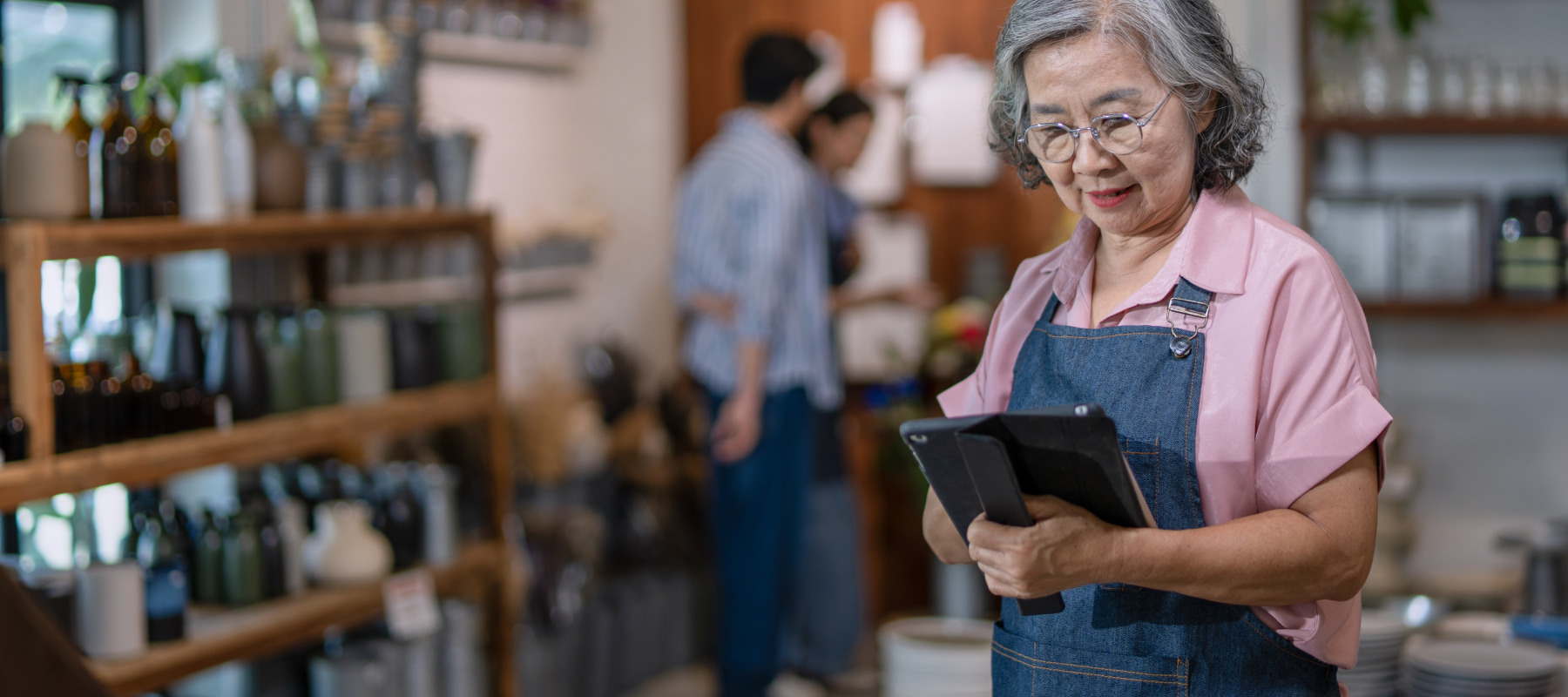 n adult woman with grey, wavy hair and glasses uses a tablet while wearing an apron.