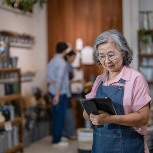 n adult woman with grey, wavy hair and glasses uses a tablet while wearing an apron.
