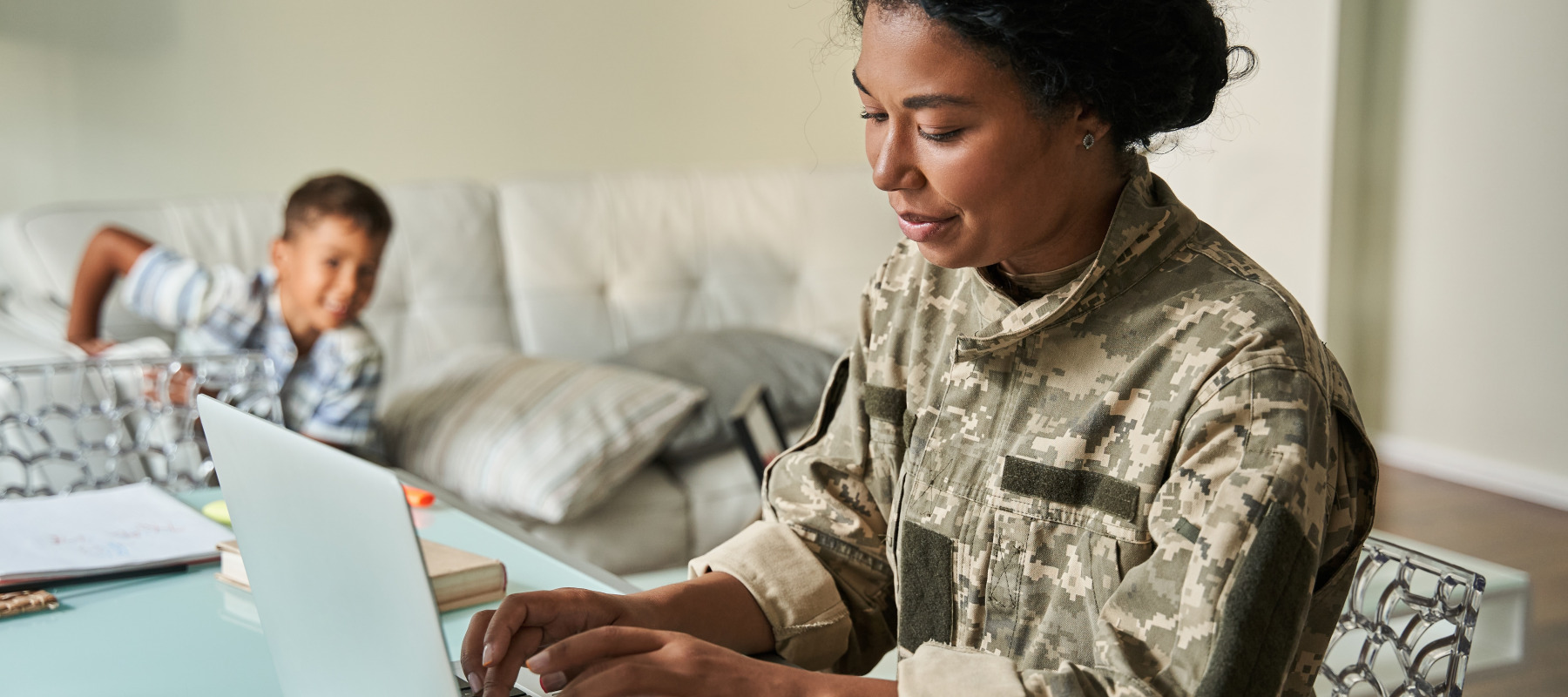 Army veteran woman working on laptop with son playing in the background