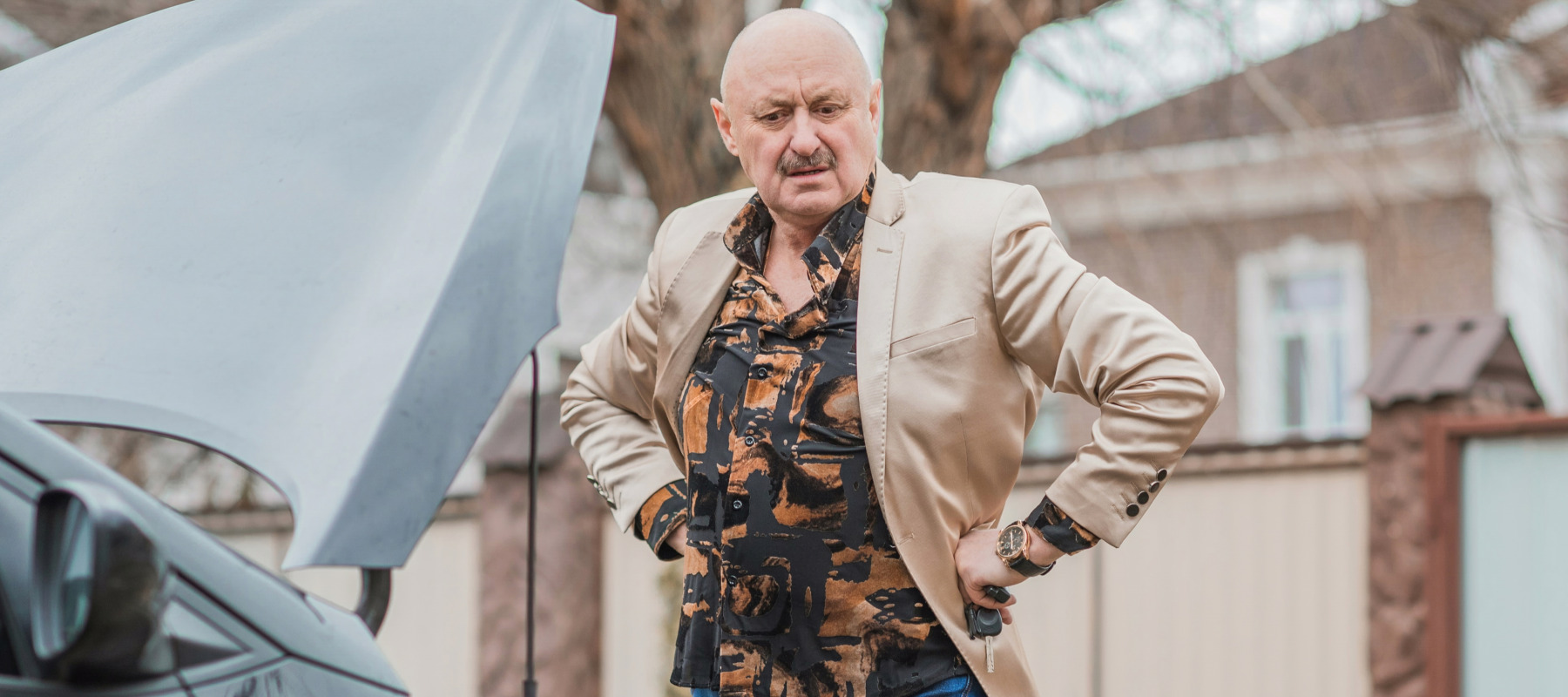 A man in a colorful shirt puts both hands on his hips with the hood of his car popped open, thinking.