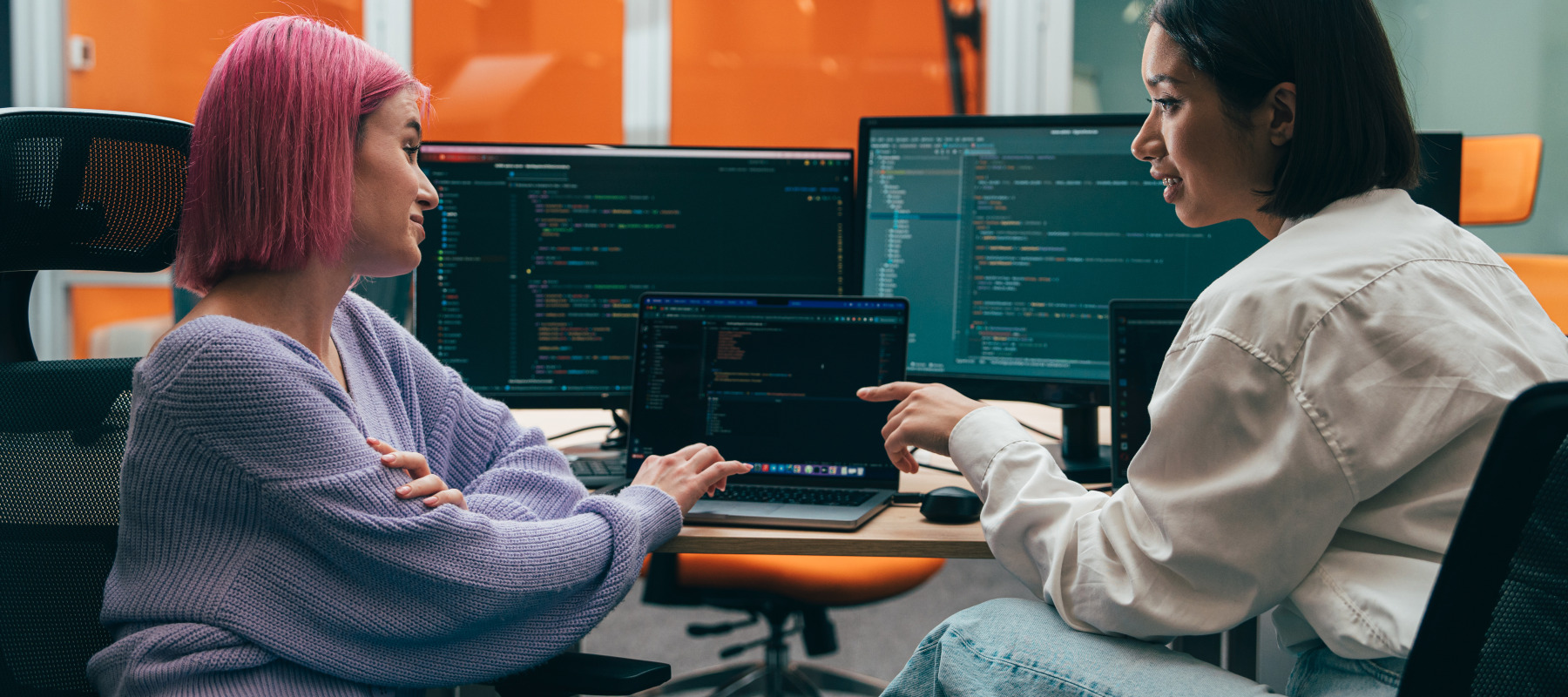 Two young adults are sitting in an office, looking at a laptop displaying code.