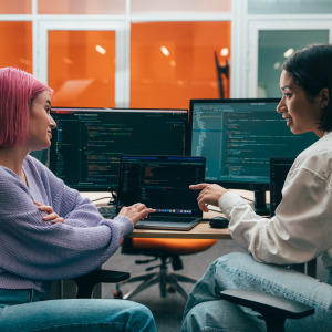 Two young adults are sitting in an office, looking at a laptop displaying code.