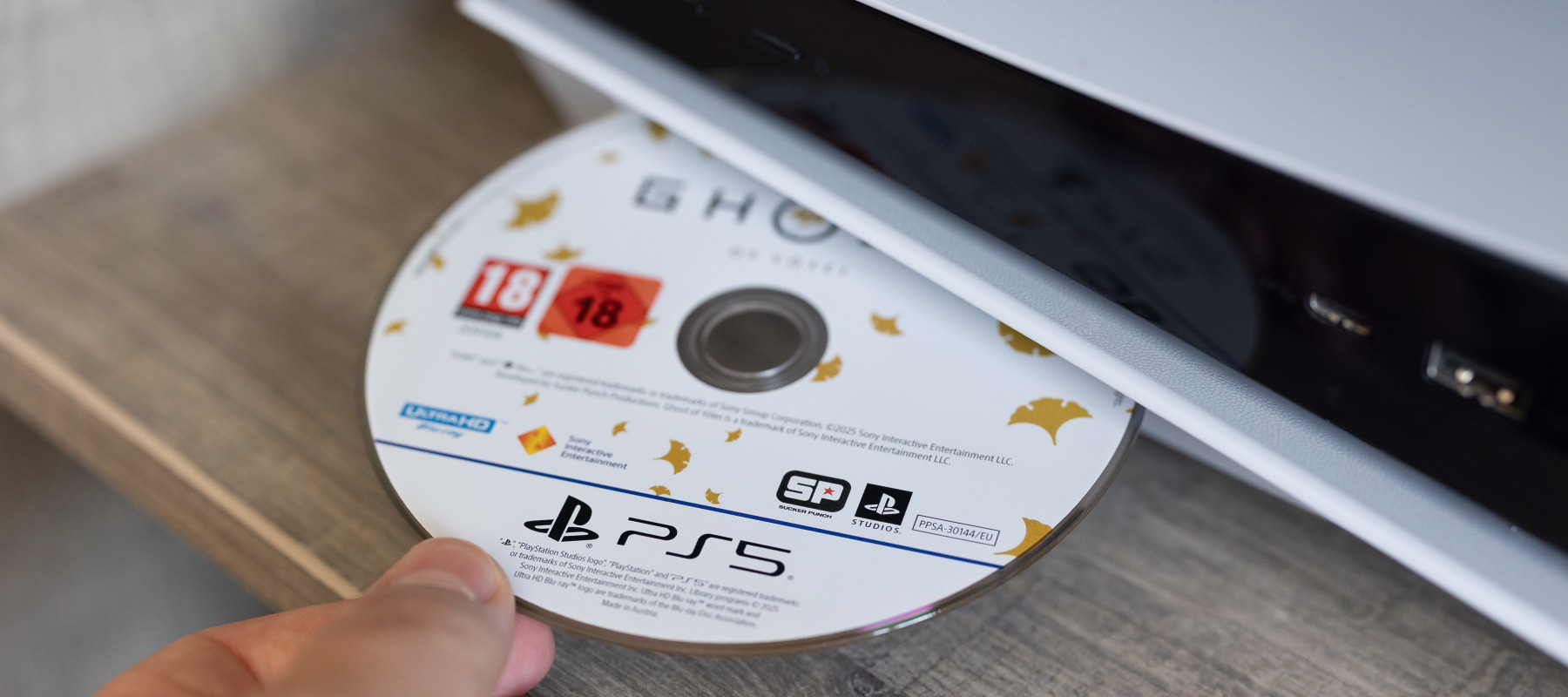 A close up image of a person's hand inserting Playstation exclusive game Ghost of Tsushima into a Playstation 5's disc port.