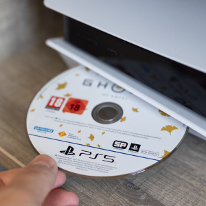 A close up image of a person's hand inserting Playstation exclusive game Ghost of Tsushima into a Playstation 5's disc port.