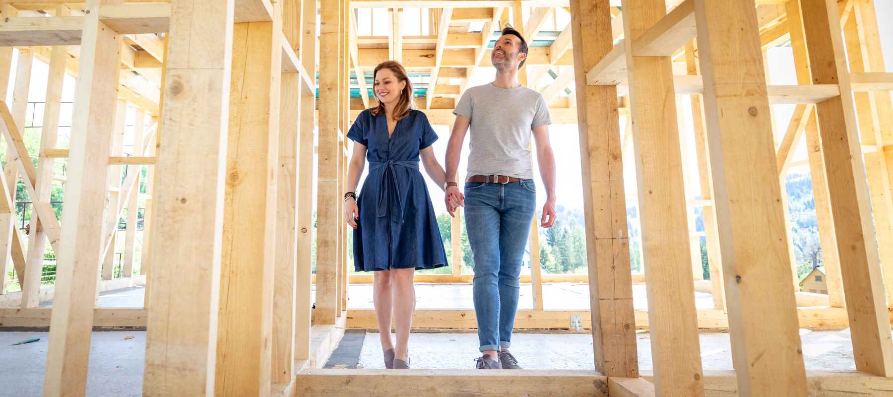A woman and a man, holding hands, stand inside the wooden framework of a house under construction.