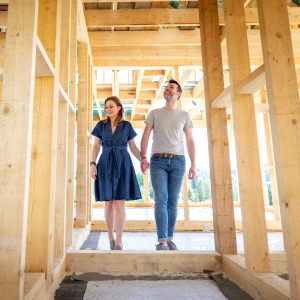 A woman and a man, holding hands, stand inside the wooden framework of a house under construction.