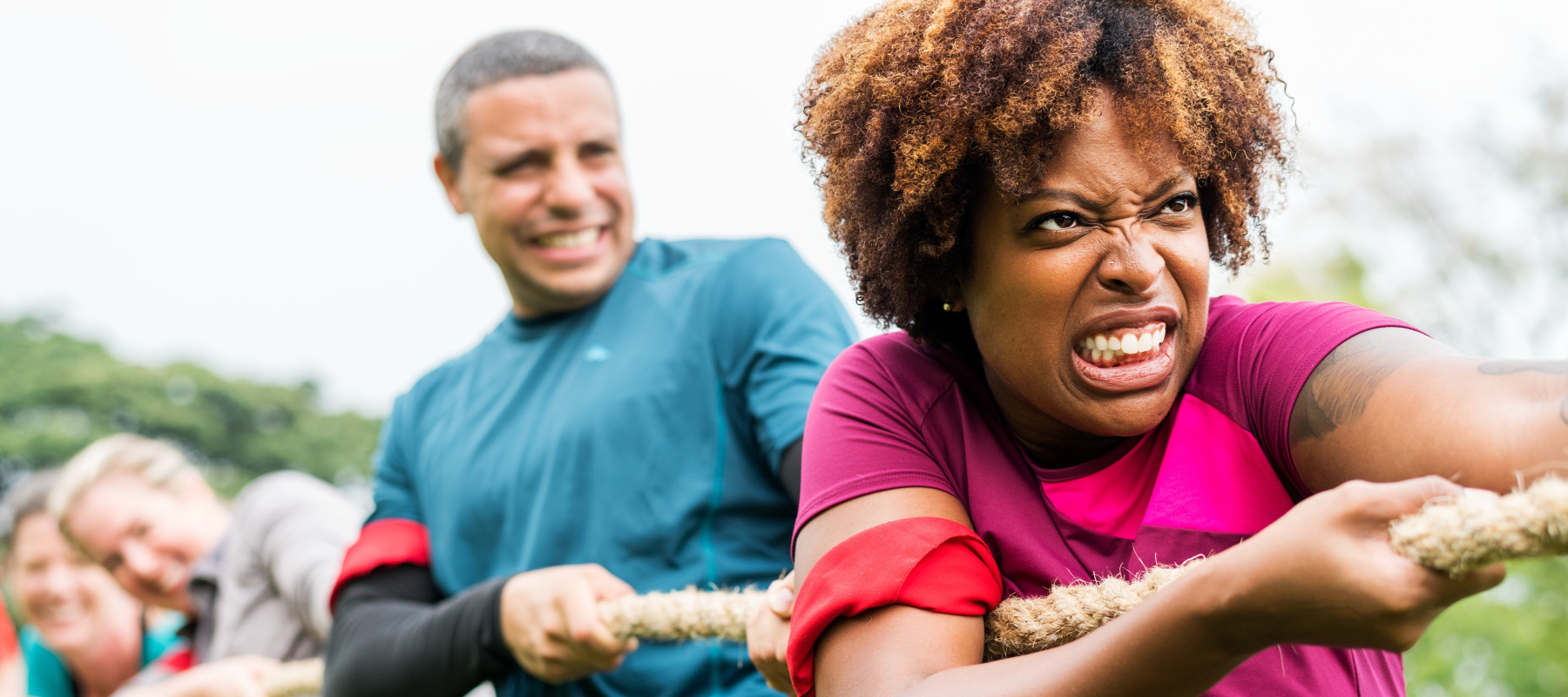 Woman grimaces as she pulls on rope in game of tug of war, with her teammates seen behind her.