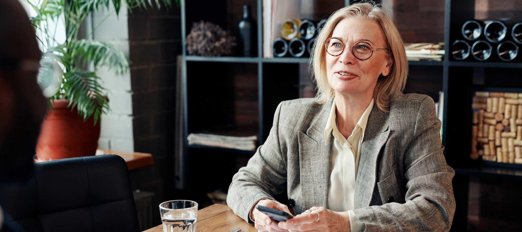 A well-dressed woman with blonde hair sits at a wooden table holding a phone and folder while having a conversation with another person.
