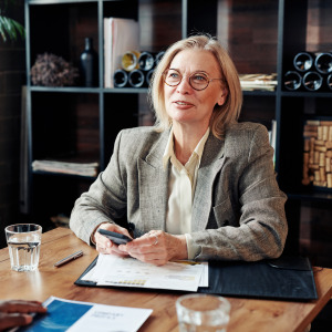 A well-dressed woman with blonde hair sits at a wooden table holding a phone and folder while having a conversation with another person.