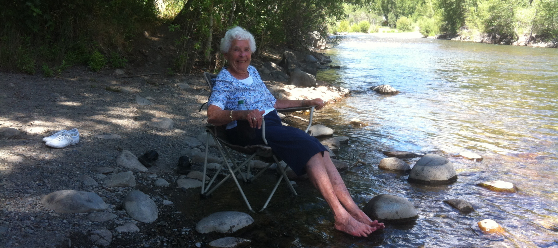 Older woman smiles, sitting in a camp chair as she dips her feet in a river.