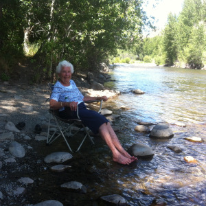 Older woman smiles, sitting in a camp chair as she dips her feet in a river.