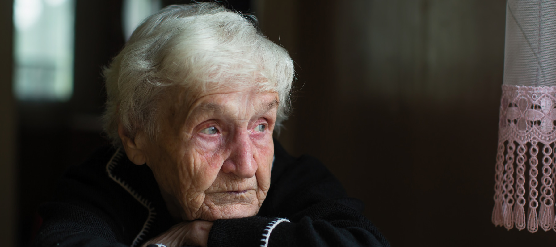 An older woman rest her chin on both wrists and looks into the middle distance with sad blue eyes.
