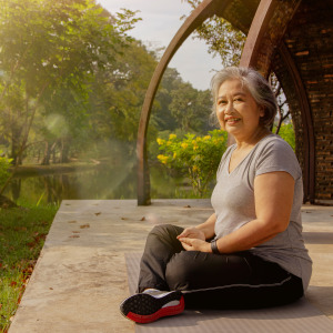 Older Asian woman sits cross-legged in the park, smiling at the camera.