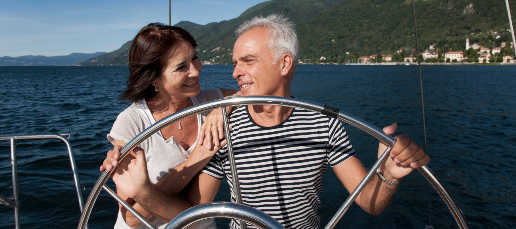 Happy couple look at each other smiling while standing behind the wheel of a sailboat.