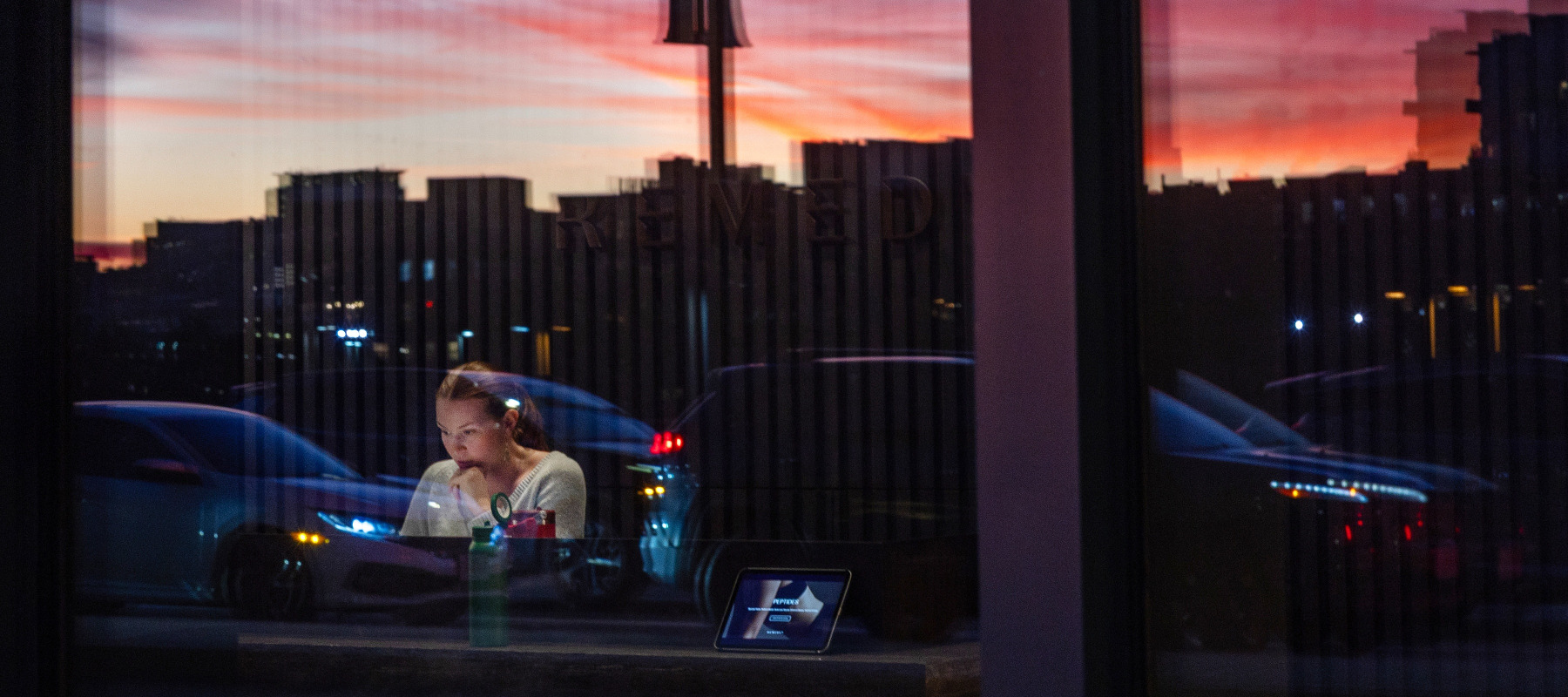 Woman seen sitting in an office at street-level, reflection of setting sun against the glass as she looks closely at her computer.