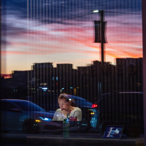 Woman seen working at a computer at street level, with reflection of sunset seen on the glass of her office.