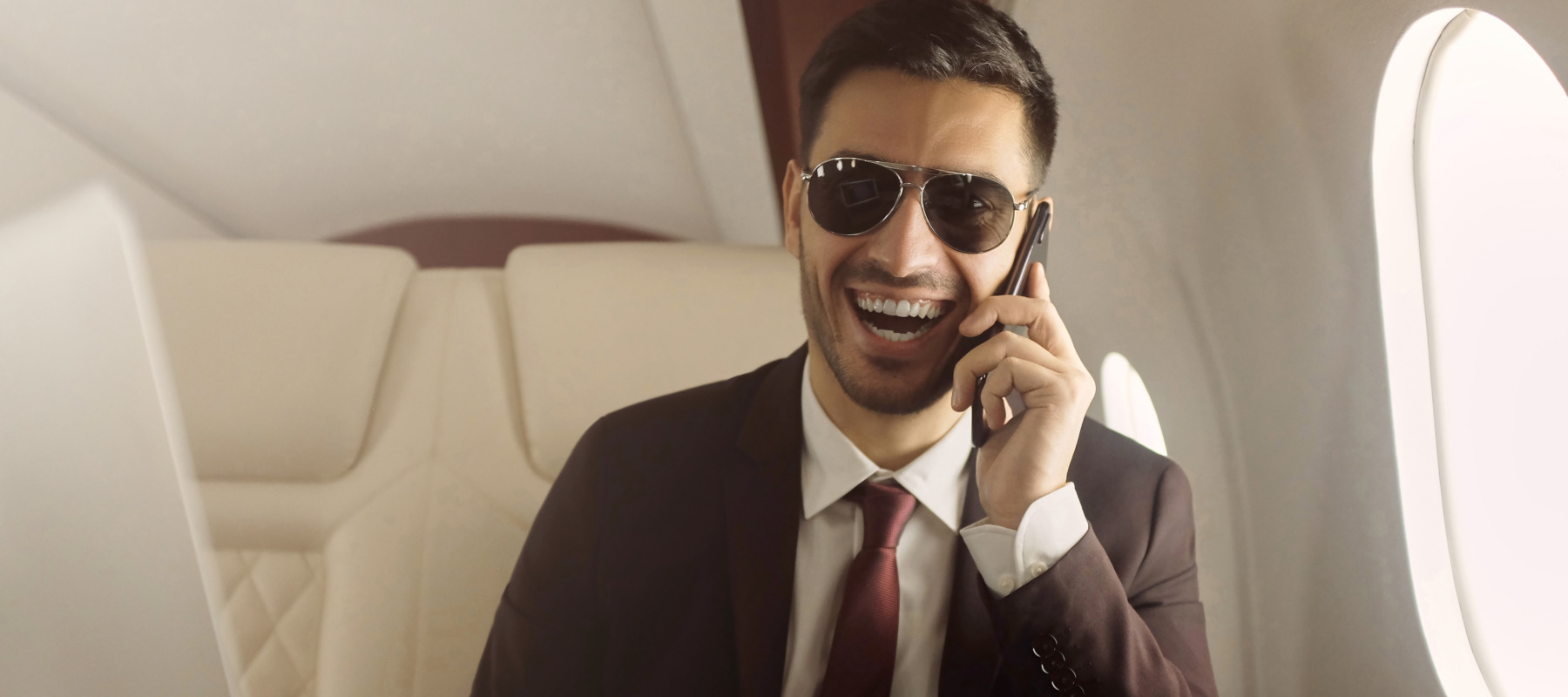 Happy laughing rich man wearing suit and sunglasses, flying in private jet, sitting at table covered with bands of cash.