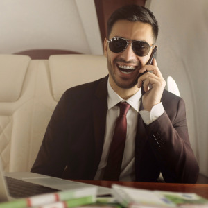 Happy laughing rich man wearing suit and sunglasses, flying in private jet, sitting at table covered with bands of cash.