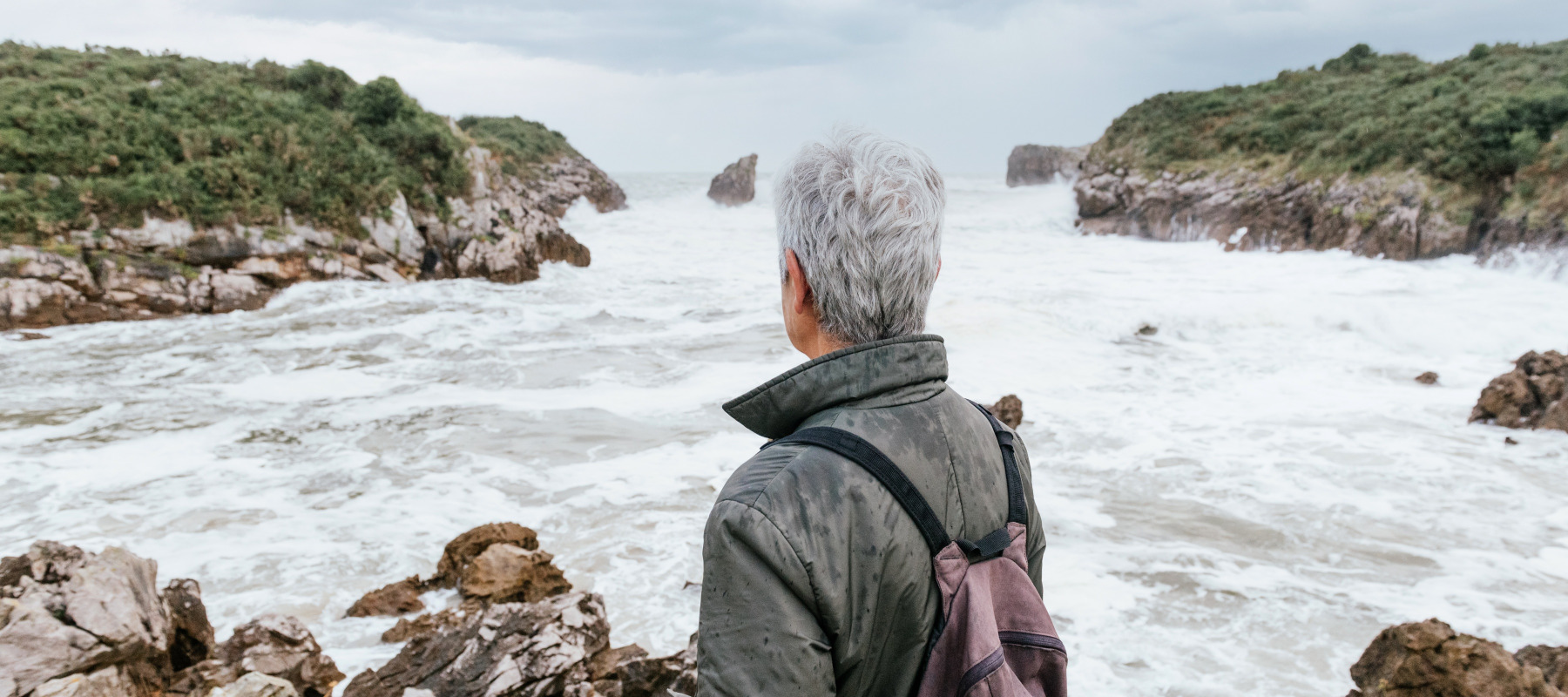Older person with grey hair seen from behind, standing in front of beach as waves crash at the shore.