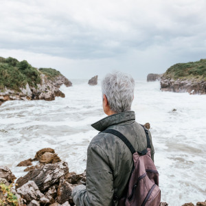 Older person with grey hair seen with back to camera, facing out at beach as waves crash on the shore.
