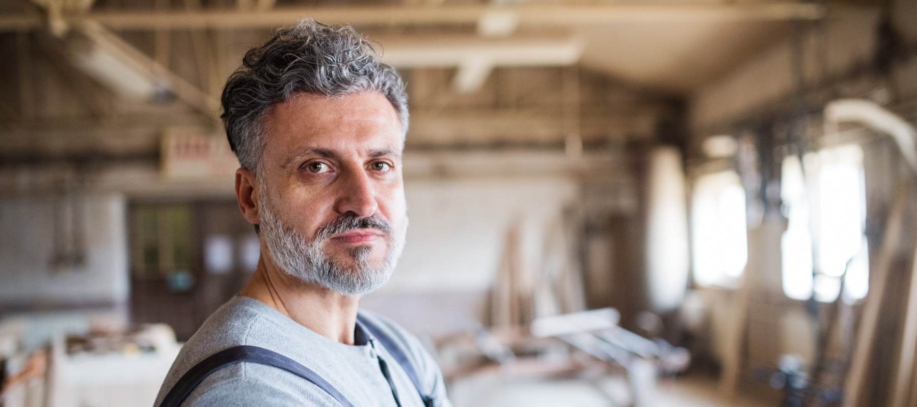 Man working in a carpentry shop looks directly at the camera, with a serious expression on his face.